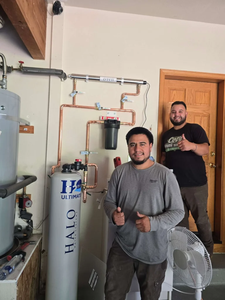 Photo Gallery: Two men stand in front of a newly installed water filtration system, both giving thumbs up. Pipes and equipment are visible on the wall next to them in a utility room.