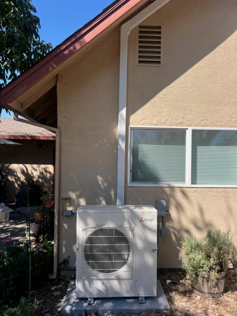 A heat pump unit is installed outside a beige house, beneath a window, with conduit running up the wall to the attic vent. Shrubs and patio furniture are nearby, creating an inviting scene worthy of a Photo Gallery.