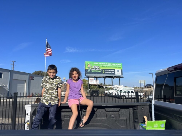 Photo Gallery: Two children sit in the bed of a pickup truck in a parking lot on a sunny day, with a U.S. flag and billboard visible in the background.