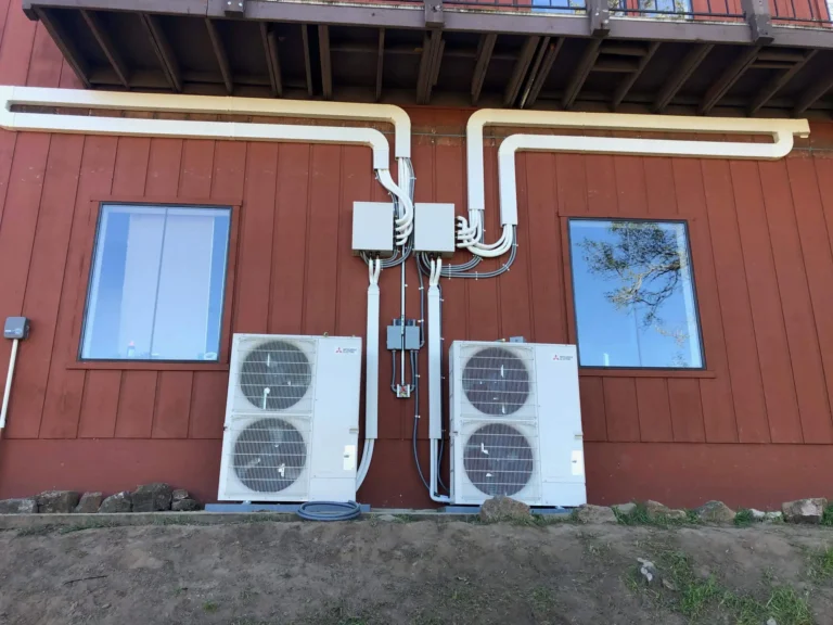 Photo Gallery: Two outdoor HVAC units with connected conduit pipes are mounted on the ground against a red building wall between two windows.