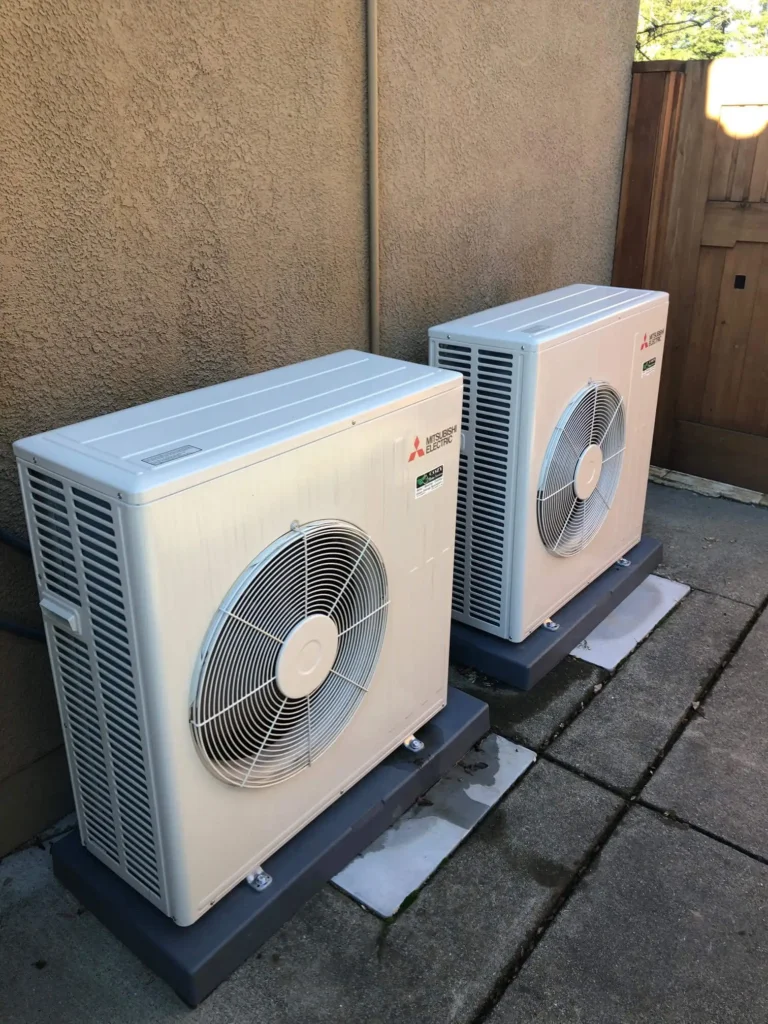 Photo Gallery: Two outdoor Mitsubishi air conditioning units are installed side by side on raised platforms outside a building, with a concrete pathway and wooden fence nearby.