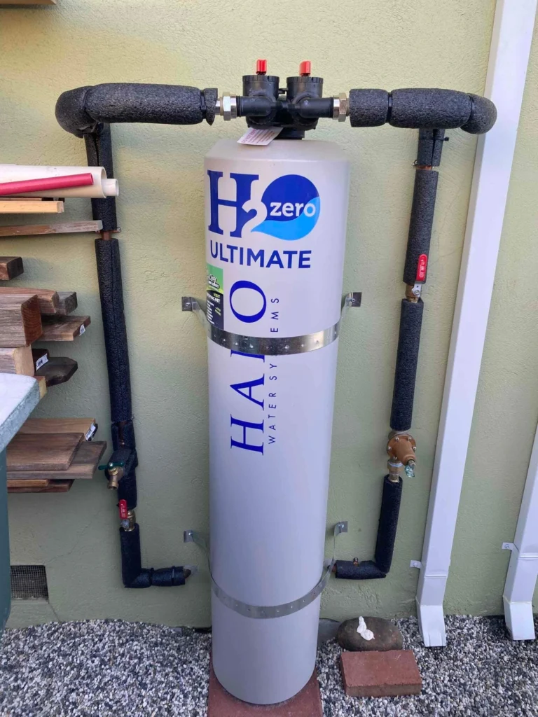 A large cylindrical water filtration system labeled "H2O ULTIMATE HALO WATER SYSTEMS" is installed outdoors with black insulated pipes connected at the top, making an impressive addition to any Photo Gallery showcasing advanced home water solutions.