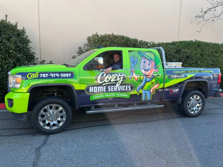 Photo Gallery: Green Cozy Home Services truck with company branding and contact details on the side; person in driver’s seat, parked on a paved surface near bushes and a wall.