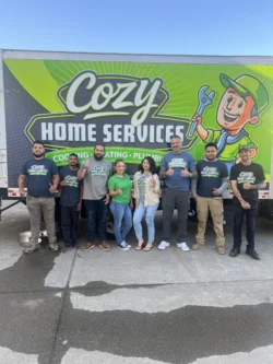 Eight people stand in front of a Cozy Home Services truck, posing for a group photo and giving thumbs up. The truck advertises cooling, heating, and plumbing services.