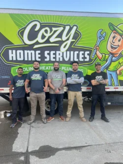 Five men standing in front of a Cozy Home Services truck, posing for a group photo. The truck features plumbing, heating, and cooling services graphics.