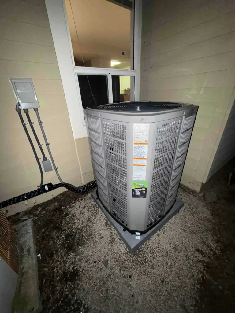 A central air conditioning unit is installed outside a house, positioned on a concrete pad beneath a window, with electrical wiring connected to a nearby wall box—perfect for featuring in your home improvement Photo Gallery.