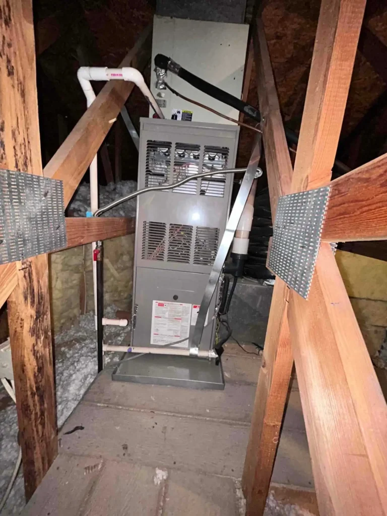 A gray HVAC furnace unit installed in an attic, surrounded by wooden beams and insulation, with visible pipes and ductwork attached—perfect for showcasing in a Photo Gallery.