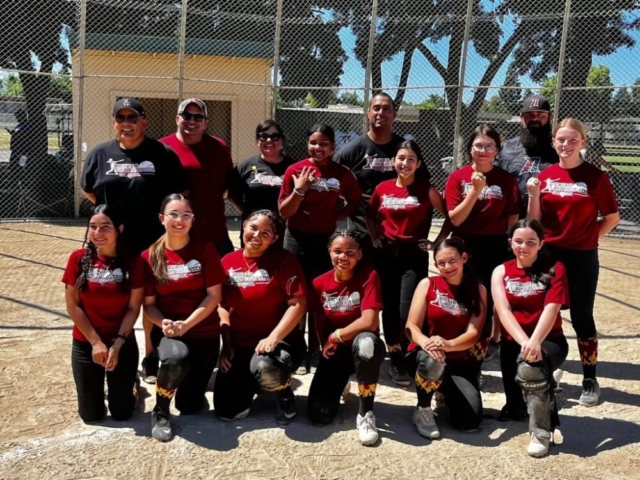 A girls' softball team and their coaches pose together on a dirt field in front of a chain-link fence, all wearing matching maroon shirts and smiling at the camera for the team’s photo gallery.