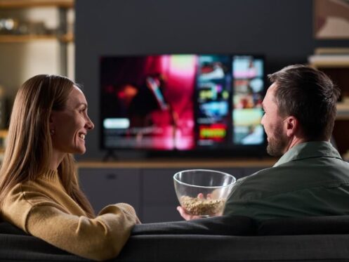 A woman and a man sit on a couch with a bowl of popcorn, watching TV with a streaming service interface displayed on the screen, enjoying perfect cooling thanks to efficient air conditioning that prevents any uneven temperature in the room.
