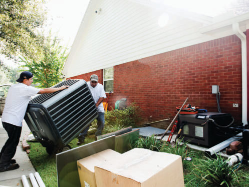 Two workers move an oversized air conditioner outside a red-brick house, with tools and equipment scattered on the lawn nearby.